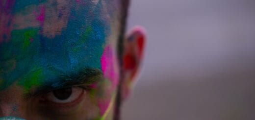 Ein weißer Mensch in einem weißen T-Shirt und mit einer Maske, der bunte Farbe im Gesicht, auf der Maske und auf dem T-Shirt hat.