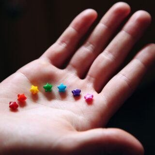 Auf einer offenen weißen Hand werden im Halbkreis bunte Sternchen gehalten in Regenbogenfarben.
